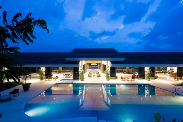 The image shows a luxurious building with an illuminated swimming pool at twilight, surrounded by contemporary furnishings and open spaces.