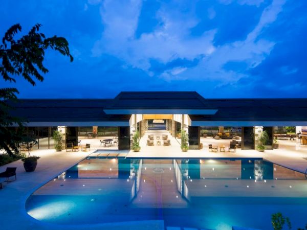 The image shows a luxurious building with an illuminated swimming pool at twilight, surrounded by contemporary furnishings and open spaces.
