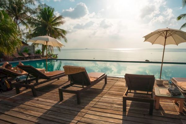 A serene poolside scene with lounge chairs, umbrellas, palm trees, and a stunning view of the ocean under a bright sky, creating a relaxing atmosphere.