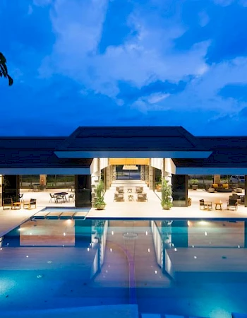 A luxurious, modern building with an illuminated swimming pool at dusk, featuring open lounge areas and surrounded by greenery and a dramatic sky.