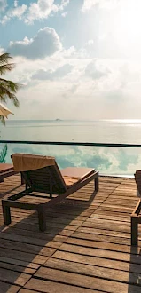 Poolside deck with loungers, umbrellas, palm trees, and a serene view of a tranquil ocean under a partly cloudy sky with sunlight.