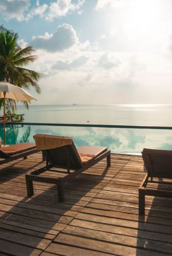 A serene poolside scene with lounge chairs, umbrellas, and palm trees overlooking a calm sea under a partly cloudy sky as the sun shines brightly.