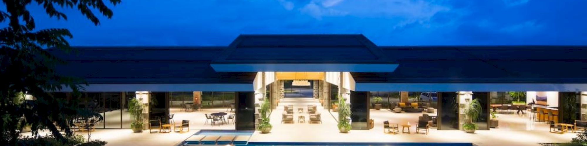 A luxurious building with a large, illuminated swimming pool in front, surrounded by lounge chairs and landscaped greenery under a twilight sky.