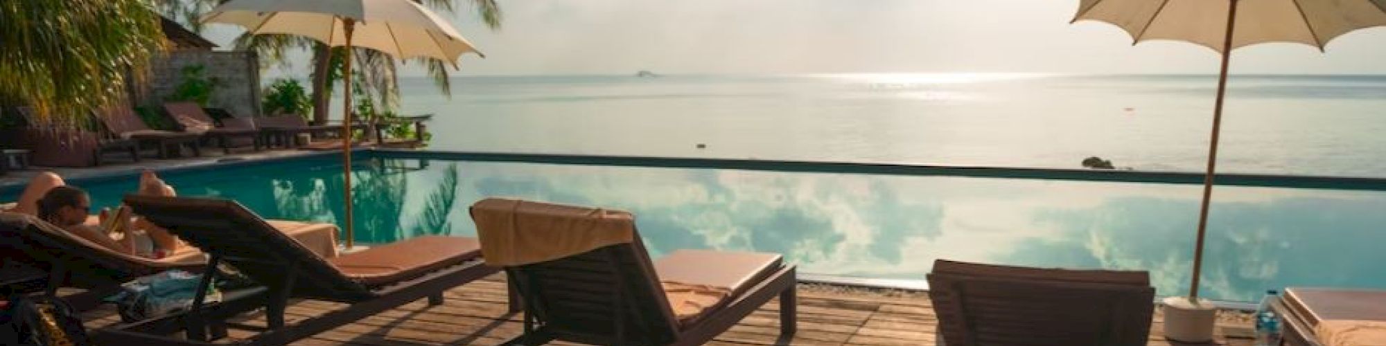 Lounge chairs and umbrellas by a pool overlooking the ocean, with palm trees and a bright, sunny sky in the background.