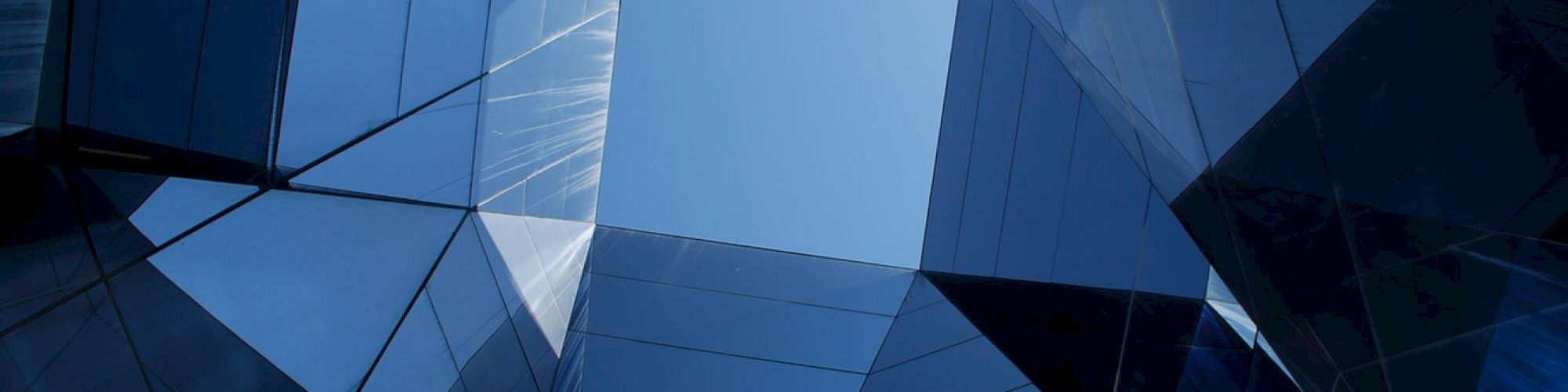The image shows a geometric arrangement of glass buildings reflecting light and creating an abstract appearance, with the blue sky visible through the center.