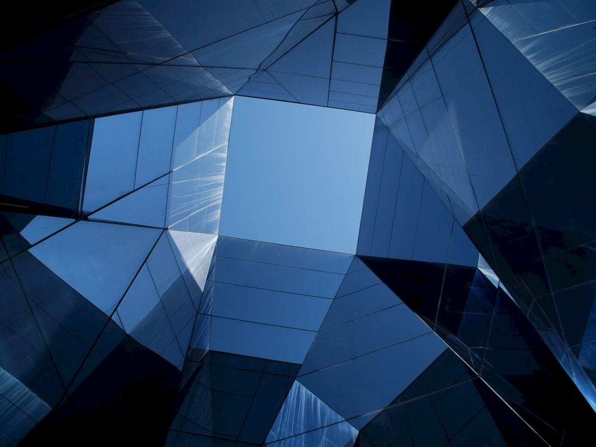 The image shows an architectural view looking up at a geometric arrangement of reflective glass panels with the sky visible in the center.
