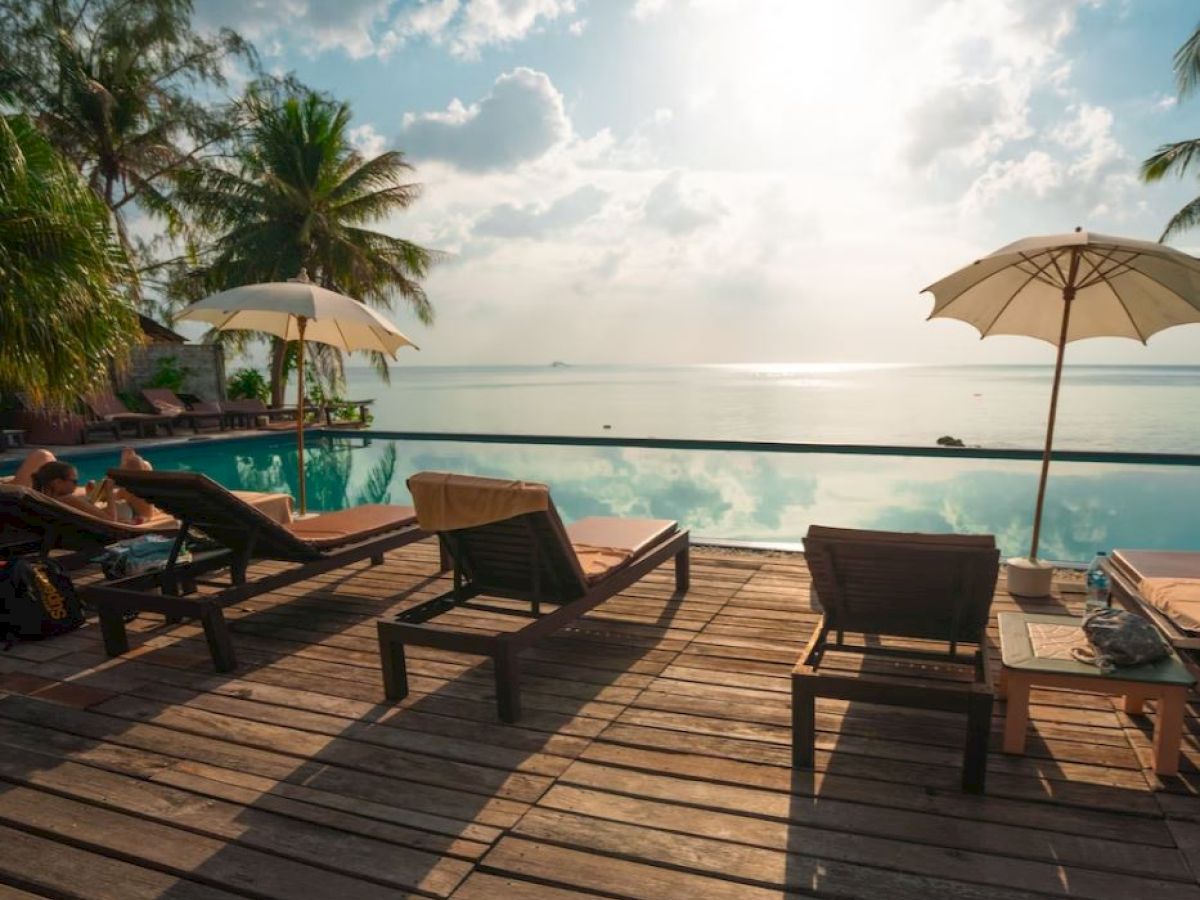 The image depicts a serene poolside scene with lounge chairs, umbrellas, and a view of the ocean, under a partly cloudy sky and surrounded by palm trees.
