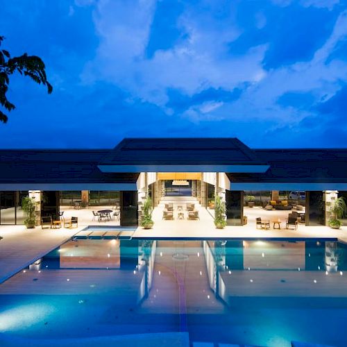 The image shows a luxurious, modern building with an illuminated outdoor swimming pool against a backdrop of a blue sky.