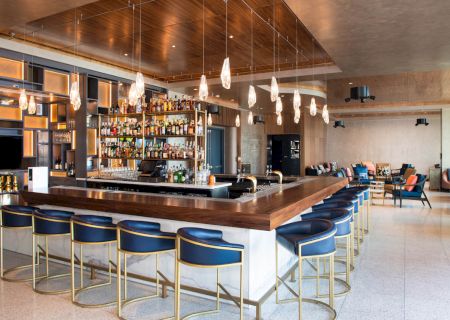 The image shows a modern bar with blue barstools, hanging lights, a wide selection of drinks, and comfortable seating in the background.