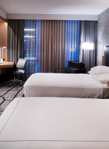 The image shows a modern hotel room with two neatly made beds, a wall-mounted TV, a desk with a chair, and stylish décor and lighting elements.