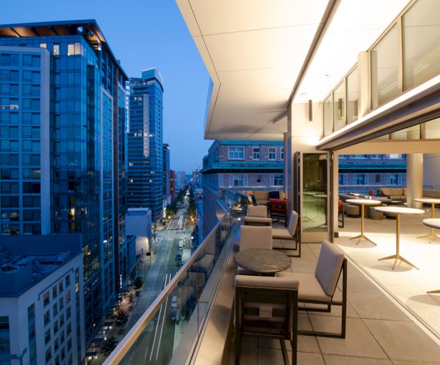A modern outdoor terrace at dusk overlooks a city with tall buildings, featuring seating areas, tables, and a view down a busy street below.