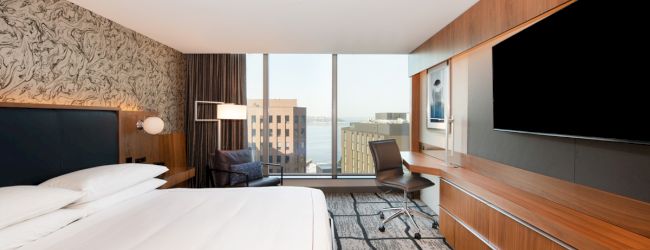 A modern hotel room with a large bed, flat-screen TV, desk, chair, and window view of a cityscape featuring tall buildings and water.