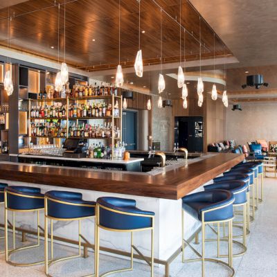 The image shows a modern bar with a wooden countertop, blue cushioned bar stools, hanging lights, and a well-stocked liquor shelf in the center.