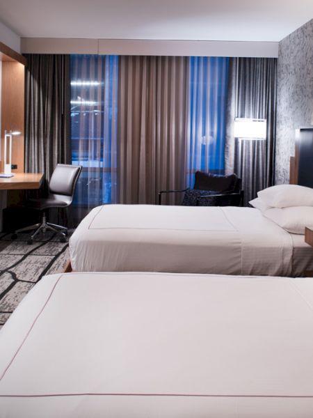 This image shows a modern hotel room with two beds, a wall-mounted TV, a desk with a chair, and large windows with curtains, with modern lighting.