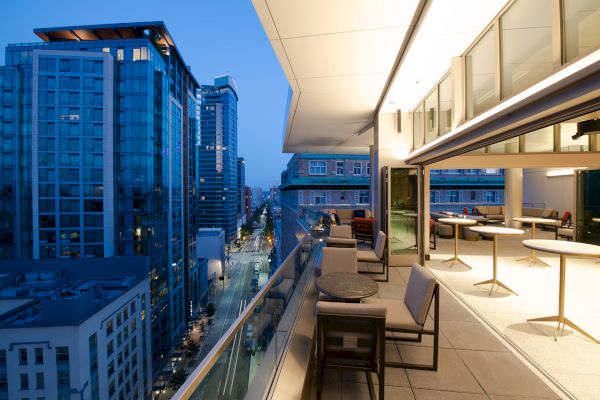 Rooftop terrace with tables and chairs overlooking a cityscape at dusk, featuring modern buildings and well-lit streets.