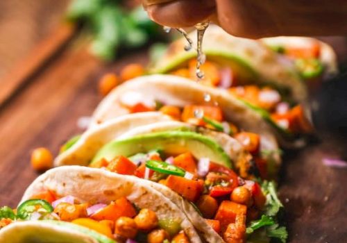 A hand squeezing lime juice onto a row of colorful, vegetable-filled tacos on a wooden surface.