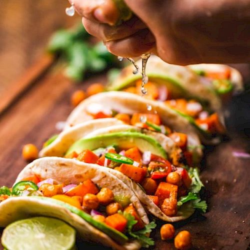 A hand squeezing lime juice onto a row of colorful, vegetable-filled tacos on a wooden surface.