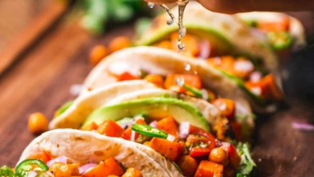 A hand squeezing lime juice onto a row of colorful, vegetable-filled tacos on a wooden surface.