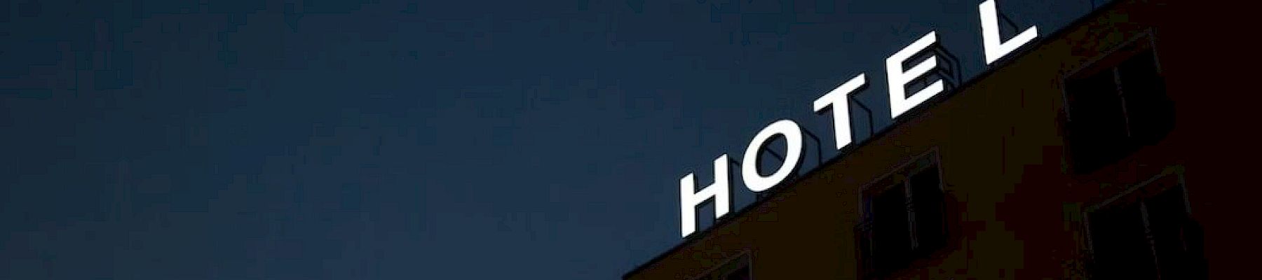 The image shows a hotel building at dusk with the illuminated sign "HOTEL" missing the letter "E".