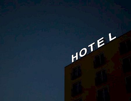 The image shows a hotel building at dusk with the illuminated sign "HOTEL" missing the letter "E".