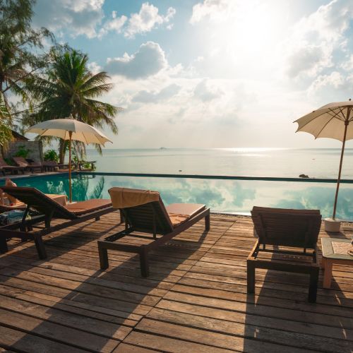 A serene ocean view with lounge chairs and umbrellas on a wooden deck by a pool, surrounded by palm trees under a partly cloudy sky at sunset.