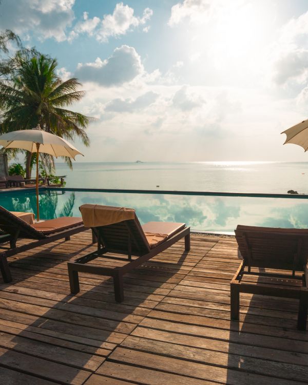 A serene ocean view with lounge chairs and umbrellas on a wooden deck by a pool, surrounded by palm trees under a partly cloudy sky at sunset.