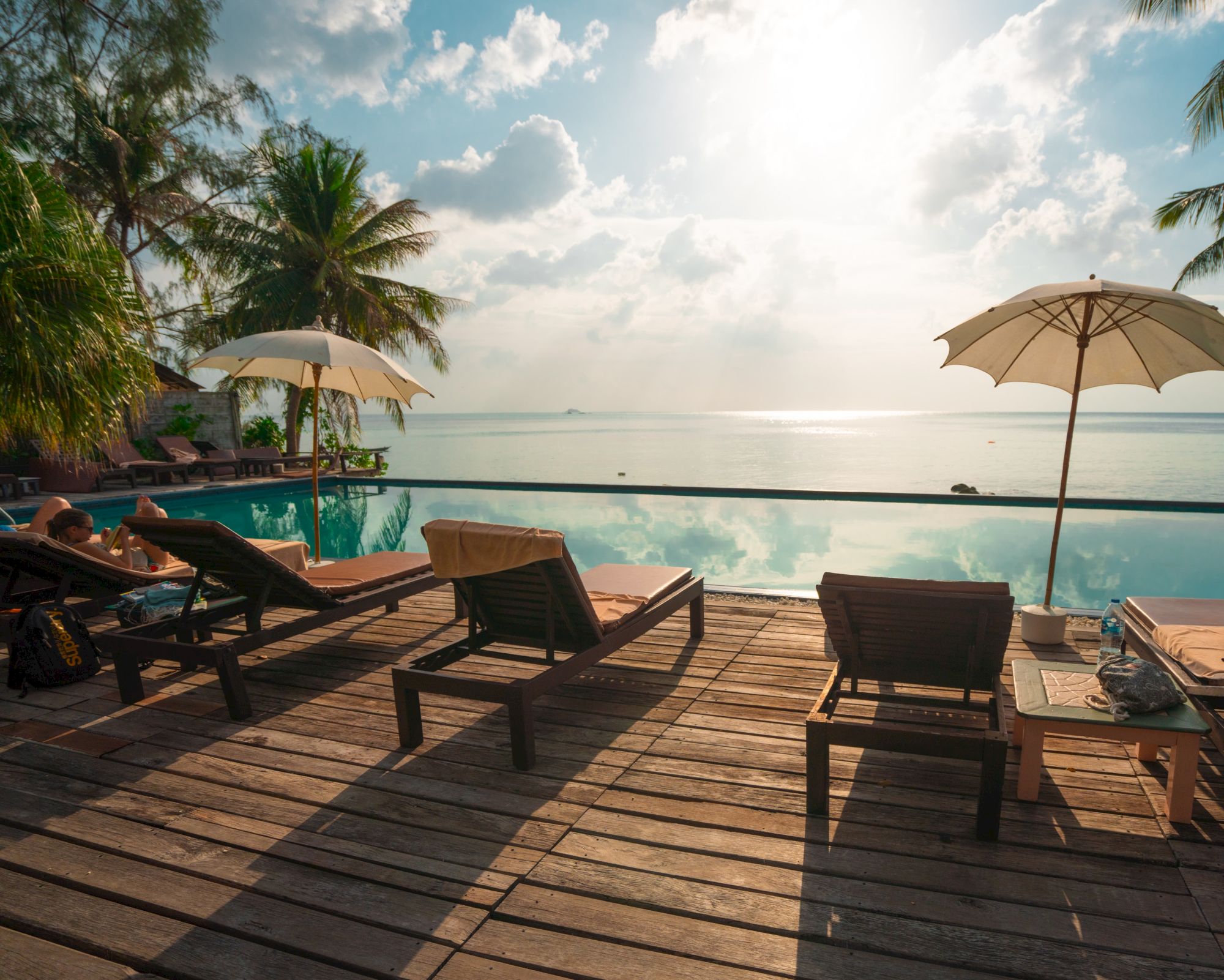 A serene beach scene with lounge chairs and umbrellas on a wooden deck beside a tranquil pool, overlooking the ocean under a sunny sky.