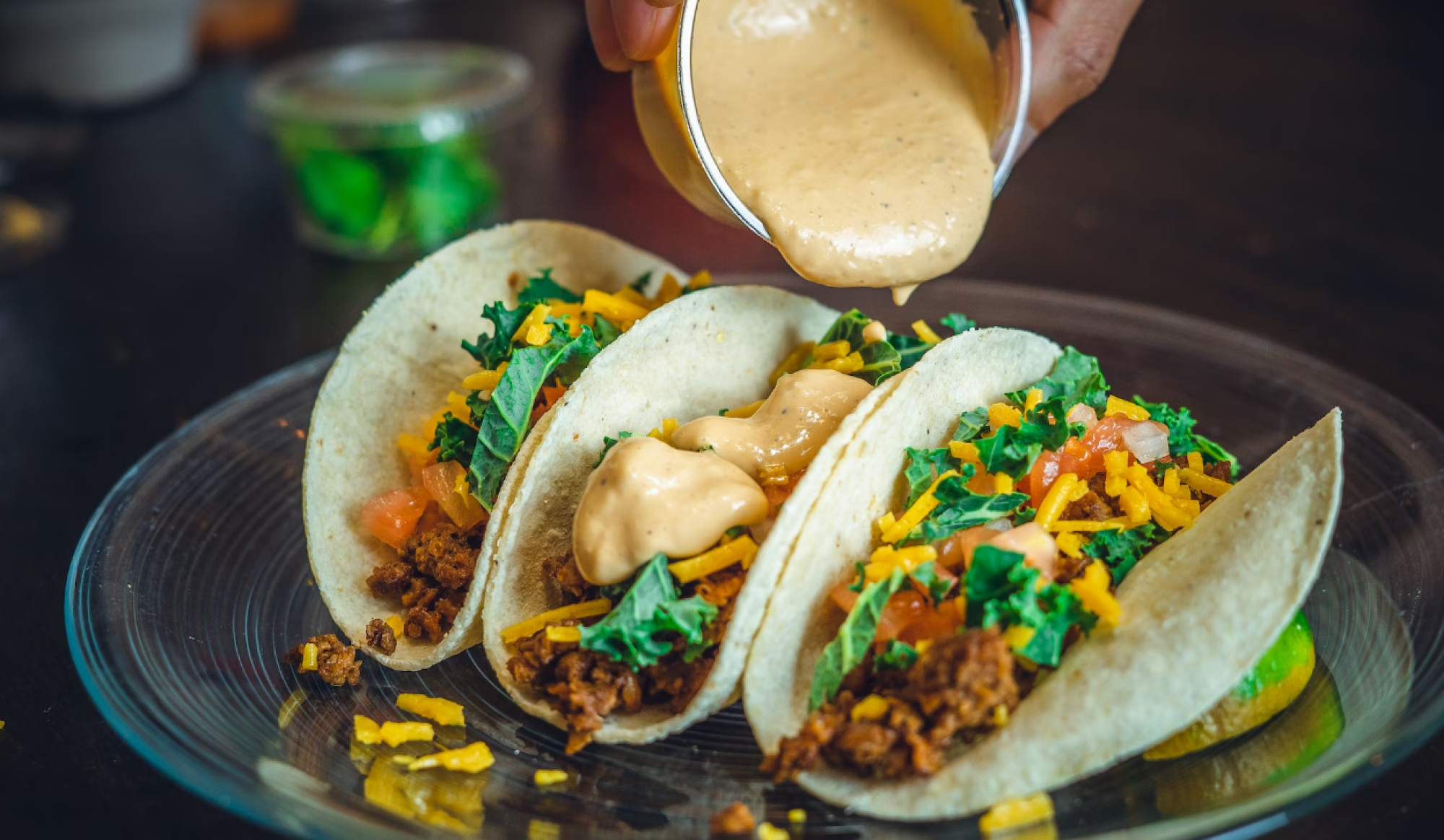 Someone is pouring creamy sauce onto a plate of three tacos filled with cheese, leafy greens, and diced tomatoes, all on soft tortillas.
