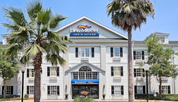 The image shows the front of a Baymont Inn & Suites hotel with palm trees, a well-maintained entrance, and the hotel’s signage.