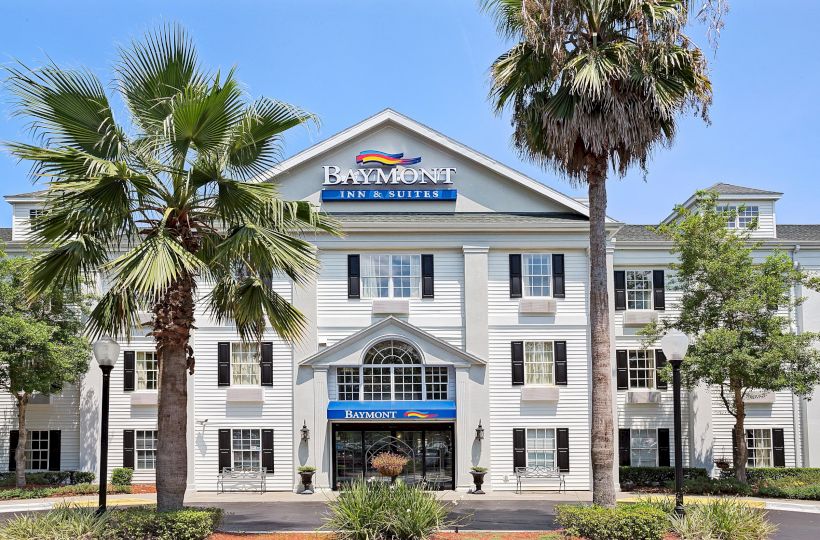 The image shows the front of a Baymont Inn & Suites hotel with palm trees, a well-maintained entrance, and the hotel’s signage.