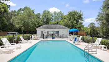 An outdoor pool surrounded by lounge chairs and tables with umbrellas, next to a small building and greenery in the background.