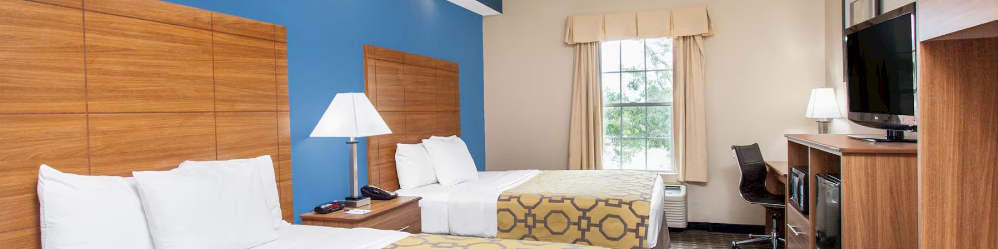 A hotel room with two double beds, lamps, a TV, a desk, and a window with curtains. The room has blue and beige walls and patterned bedspreads.