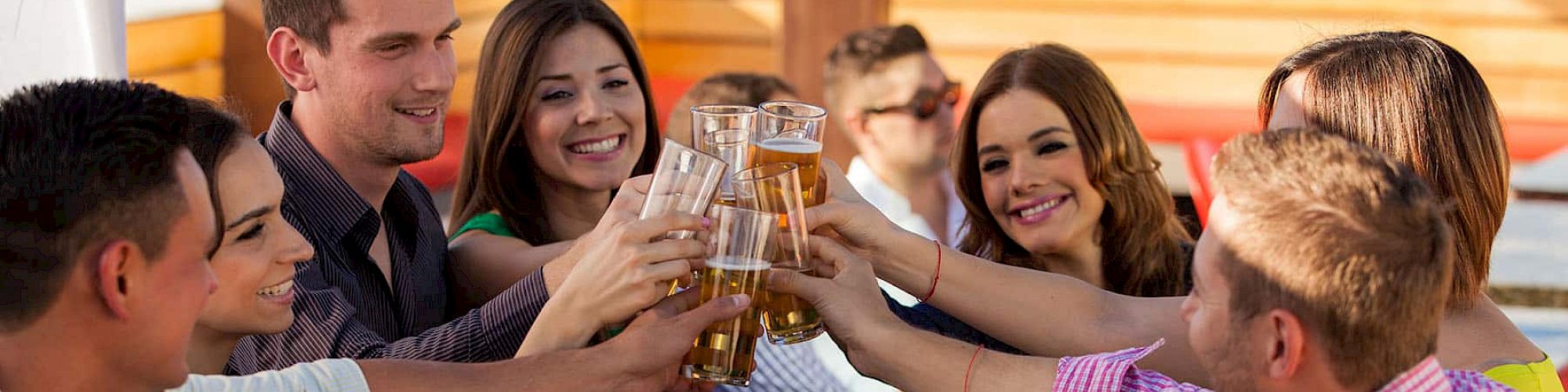 A group of people sitting around a wooden table outdoors, raising their drinks and toasting, while there is food on the table in front of them.