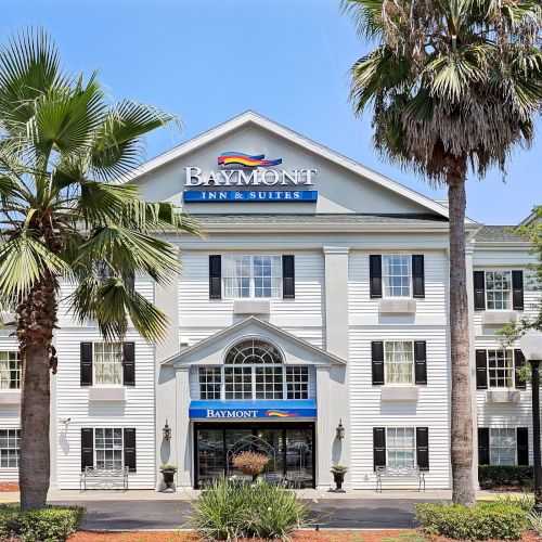 The image shows the exterior of a Baymont Inn & Suites hotel, featuring palm trees, white facade, and the hotel's signage under a clear sky.