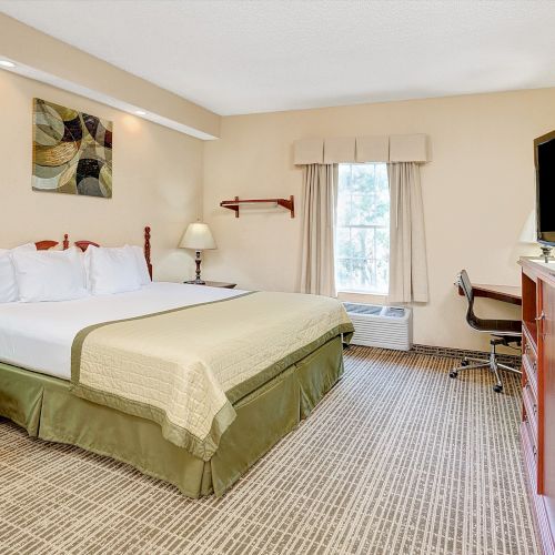 A hotel room with a king-size bed, bedside tables, lamps, a TV, a desk with a chair, a window, and a patterned carpet.