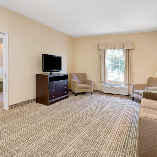 A spacious hotel room with a living area featuring a TV, sofa, armchairs, and a lamp, with a separate bedroom visible in the background through an open doorway.