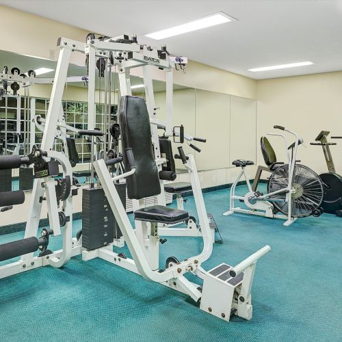 A small gym room with various exercise equipment including a multi-station machine, exercise bikes, and a treadmill, on a blue carpeted floor.