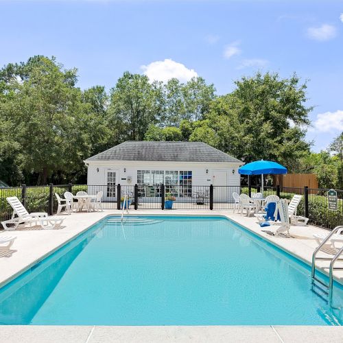 This image shows an outdoor swimming pool with lounge chairs, a blue umbrella, and a small building in the background surrounded by trees.