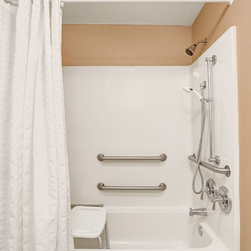 A bathroom with a white bathtub, handrails, a showerhead, and a white shower curtain in a room with tan walls and a ceiling light fixture.