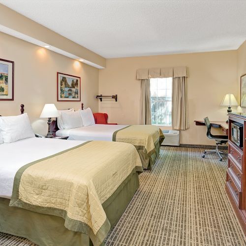 A hotel room with two beds, with beige and green bedding, TV on a stand, desk and chair by a window, and framed artwork on the walls.