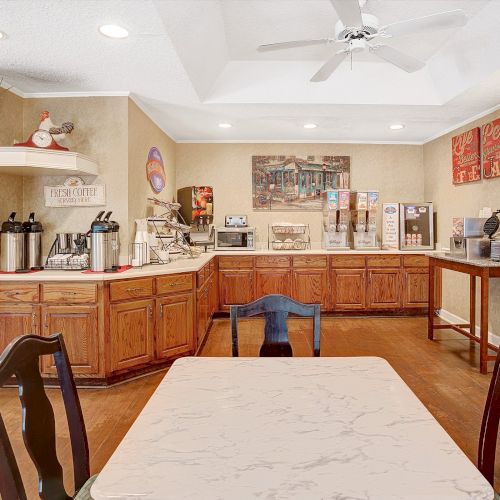 The image shows an empty, clean dining area with wooden cabinetry, beverage dispensers, a marble table, and chairs, under a ceiling fan and lights.