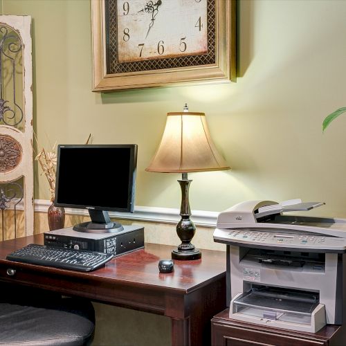 A home office setup with a wooden desk, computer, chair, table lamp, printer, plant, decorative wall panel, and wall clock above the desk.