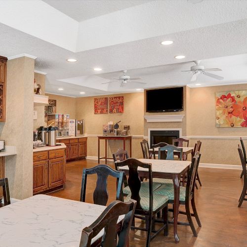 The image shows a cozy dining area with tables, chairs, a wooden counter, a coffee machine, a wall-mounted TV, and decorative artwork on the walls.