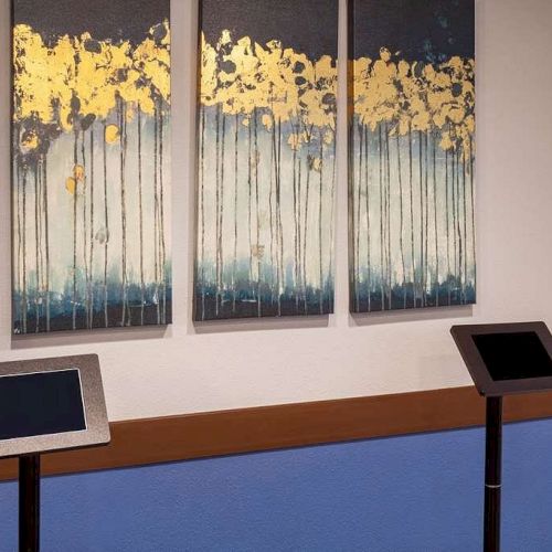 The image shows three abstract art panels on a wall, with blue and gold tones, above two black touchscreen kiosks on stands.