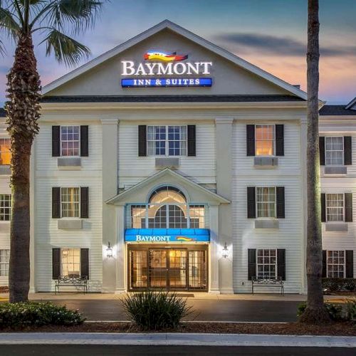 The image shows the front exterior of a Baymont Inn & Suites hotel, illuminated at night, with two street lamps and palm trees by the entrance.