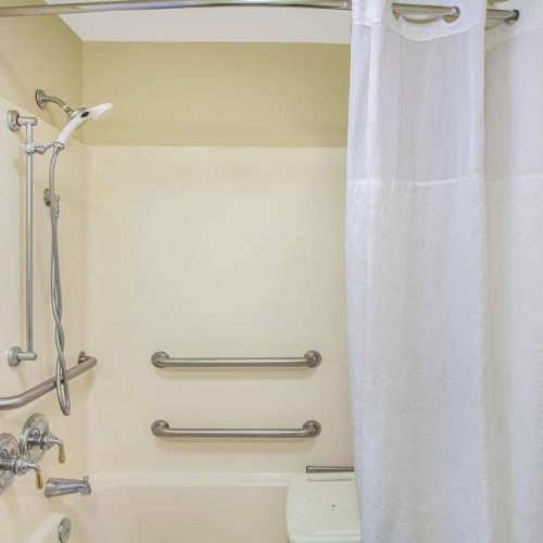 This image shows a bathroom with a tub, a showerhead, grab bars, and a white shower curtain. The wall is light-colored, and there's a toilet visible.