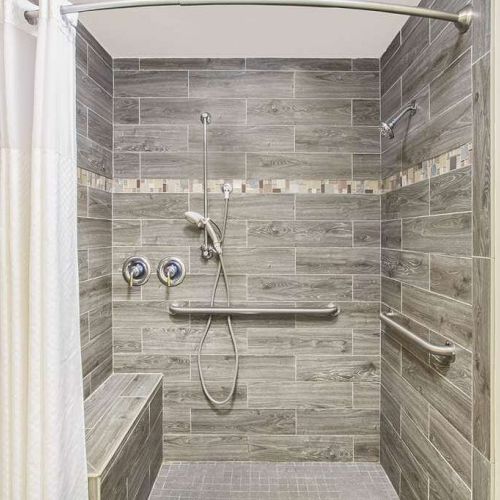 A modern shower with gray tiles, a handheld showerhead, a bench seat, grab bars, and a curtain, designed for accessibility and convenience.