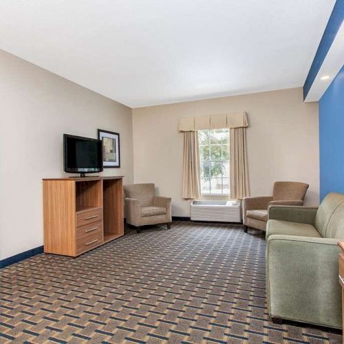 A hotel room features a seating area with a sofa, chairs, a TV on a stand, a blue accent wall, and a window with curtains.