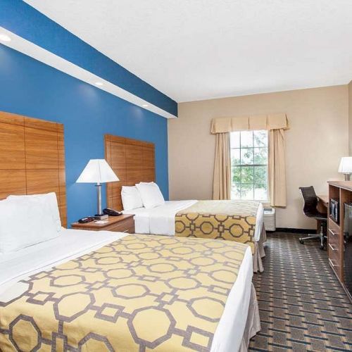 The image shows a tidy hotel room with two neatly made beds, a TV, a desk, a lamp, and a window with drapes, featuring modern furnishings.