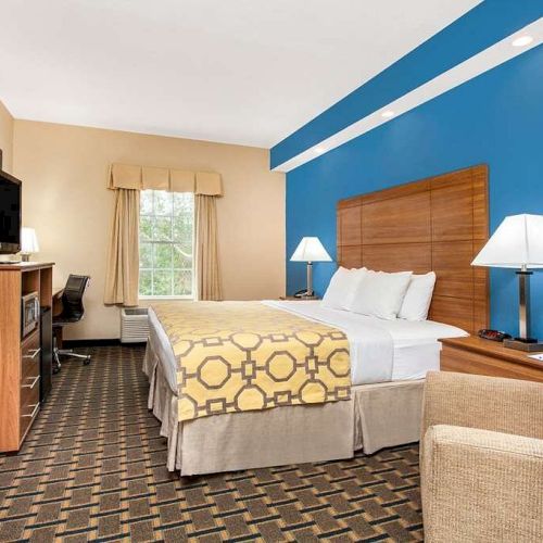 A hotel room with a bed, nightstands, lamps, a TV, a desk, and a chair; it features beige and blue walls with a window and carpeted floor.